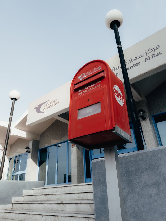 A red mailbox stands outside an Emirates Post office in Dubai, UAE, showcasing modern urban architecture.